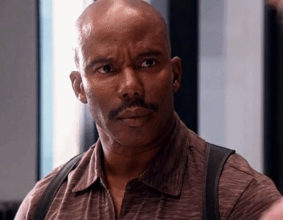 A Bald Man With A Mustache Wearing A Brown Shirt And Suspenders Looks At The Camera . GIF