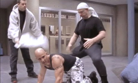 A Bald Man With A Tattoo On His Chest Is Kneeling Down GIF