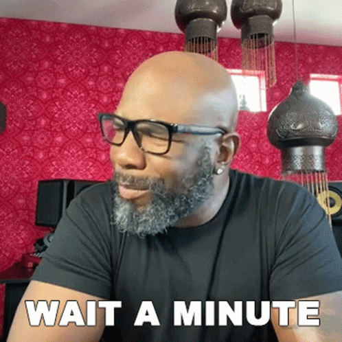 A Bald Man With Glasses And A Beard Is Sitting In Front Of A Sign That Says Wait A Minute GIF
