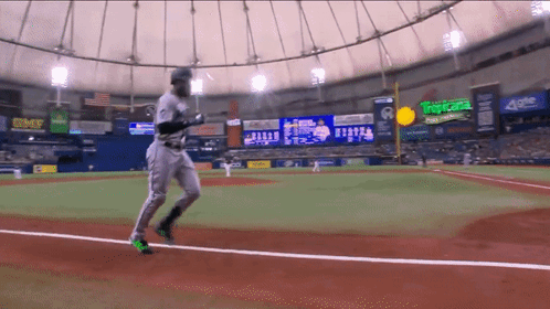 A Baseball Player Is Running On The Field In Front Of A Scoreboard That Says Tropicana On It GIF