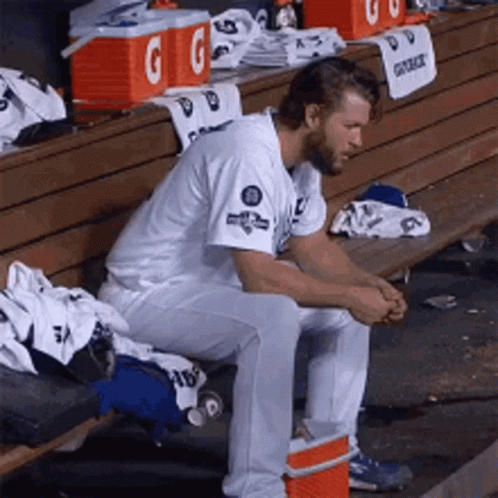 A Baseball Player Is Sitting On A Bench In A Dugout With Gatorade Coolers In The Background GIF