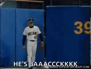 A Baseball Player Is Standing In Front Of A Blue Wall With The Words `` He 'S Baaacckkk '' Written On It . GIF