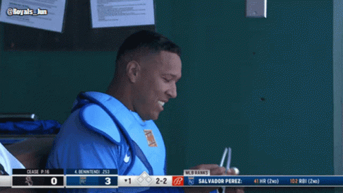 A Baseball Player Sitting In The Dugout With A Scoreboard Behind Him That Says Mlb Ranks GIF