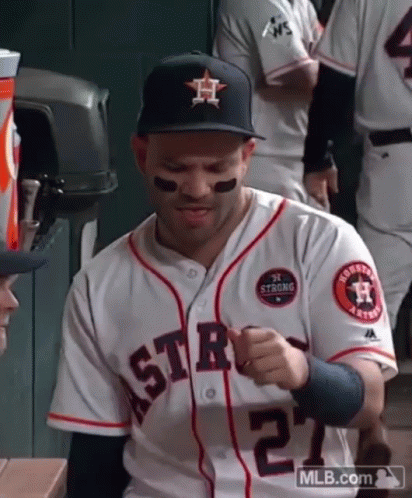 A Baseball Player Wearing A Hat And A White Jersey Is Sitting In The Dugout . GIF