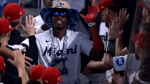 A Baseball Player Wearing A Hat With The Letter M On It Is Surrounded By His Teammates GIF