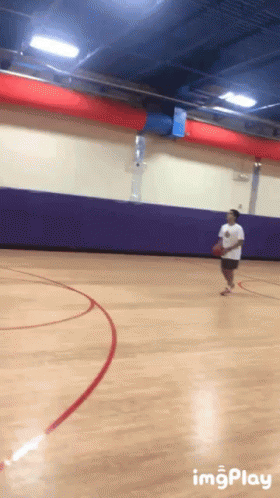 A Basketball Player Dribbles The Ball On A Court With A Purple Wall Behind Him GIF