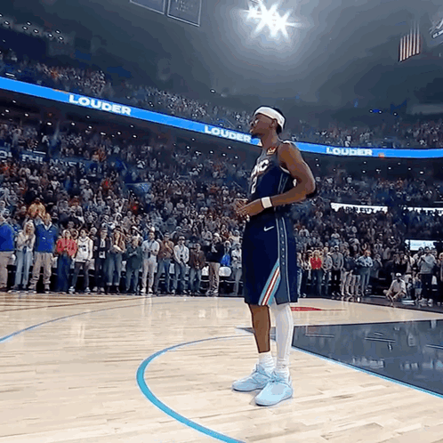 A Basketball Player Stands On The Court In Front Of A Crowd With A Banner That Says Louder On It GIF