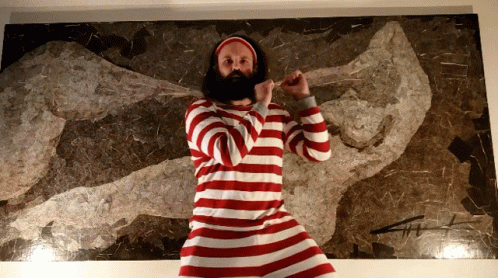 A Bearded Man In A Red And White Striped Shirt Stands In Front Of A Painting GIF