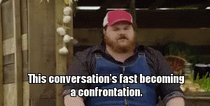 A Bearded Man Wearing Overalls And A Red Hat Is Sitting In Front Of Garlic . GIF