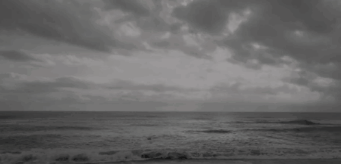 A Black And White Photo Of A Beach With A Cloudy Sky And Waves Crashing On The Shore . GIF