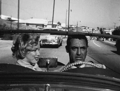 A Black And White Photo Of A Man And A Woman Driving A Car Down A Street . GIF