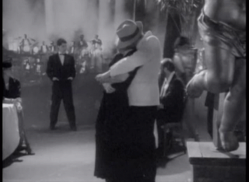 A Black And White Photo Of A Man And Woman Hugging In Front Of A Crowd GIF