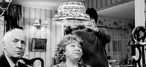 A Black And White Photo Of A Man Holding A Cake Over A Woman 'S Head In A Living Room . GIF