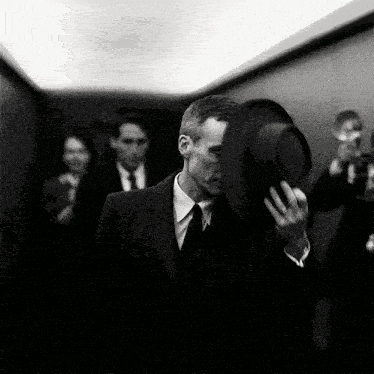 A Black And White Photo Of A Man In A Suit And Tie Holding A Hat GIF