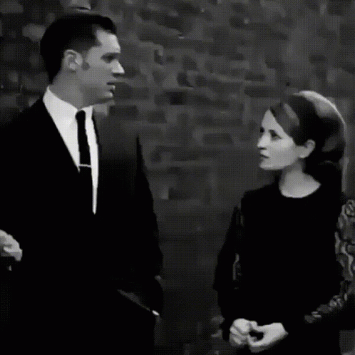 A Black And White Photo Of A Man In A Suit And Tie Standing In Front Of A Brick Wall GIF