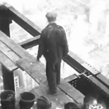 A Black And White Photo Of A Man In A Suit Standing On A Bridge . GIF