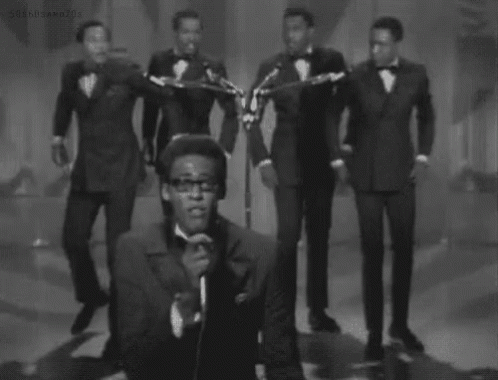 A Black And White Photo Of A Man Singing Into A Microphone While A Group Of Men Play Instruments . GIF
