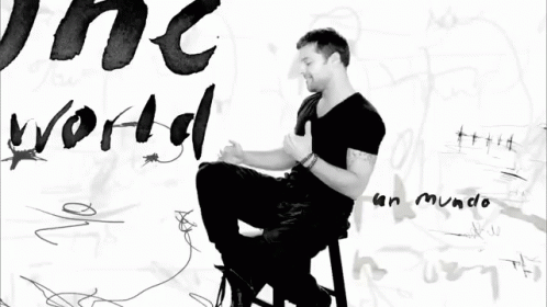 A Black And White Photo Of A Man Sitting In Front Of A Wall With The Word World Written On It GIF