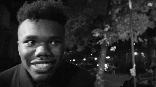 A Black And White Photo Of A Man Smiling With A Car In The Background GIF