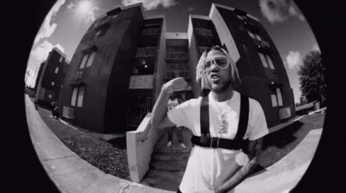 A Black And White Photo Of A Man Standing In Front Of A Building Wearing Sunglasses . GIF