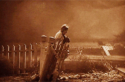 A Black And White Photo Of A Man Standing In Front Of A Fence Holding A Dog GIF