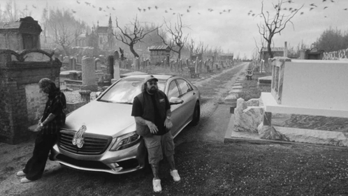 A Black And White Photo Of A Man Standing Next To A Car In A Cemetery GIF