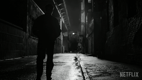 A Black And White Photo Of A Man Walking Down A Dark Alleyway With Netflix Written In The Corner GIF