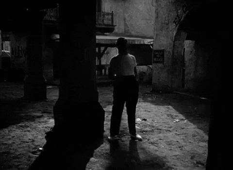 A Black And White Photo Of A Man Walking In Front Of A Sign That Says Calle Del Medio GIF