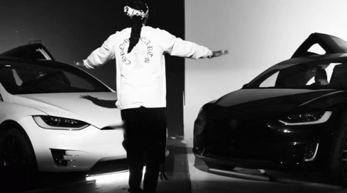 a black and white photo of a man wearing a christian dior shirt standing next to two cars