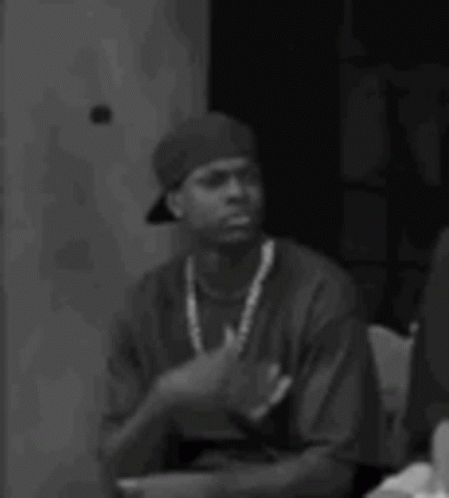 A Black And White Photo Of A Man Wearing A Hat And A Necklace Sitting Down . GIF