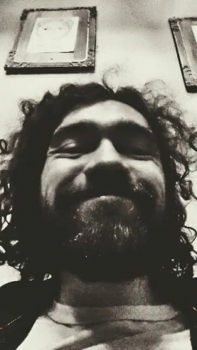 A Black And White Photo Of A Man With Curly Hair And A Beard Smiling GIF