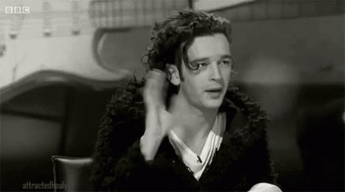 A Black And White Photo Of A Man With Curly Hair Sitting At A Table With His Hand In His Hair . GIF