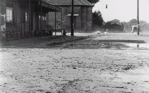 A Black And White Photo Of A Muddy Street In Front Of A Building That Says ' Warehouse ' On The Front GIF