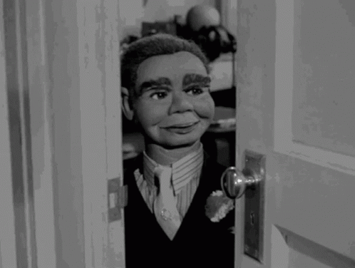 A Black And White Photo Of A Puppet In A Suit And Tie Looking Out Of A Door GIF