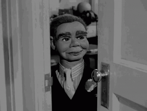 A Black And White Photo Of A Puppet Looking Out Of A Door GIF