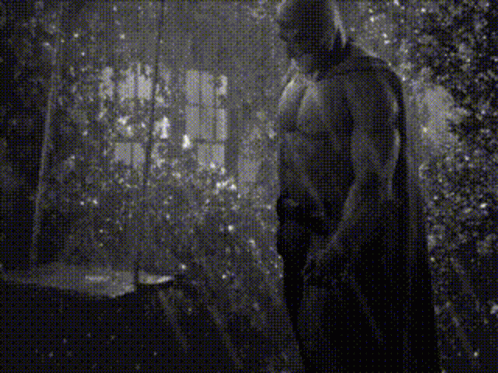 A Black And White Photo Of A Shirtless Man In A Cape Standing In A Room . GIF