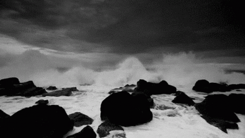 A Black And White Photo Of A Stormy Ocean With Waves Crashing Against Rocks . GIF