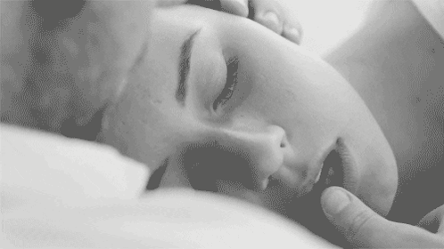 A Black And White Photo Of A Woman Laying On A Bed With Her Eyes Closed And A Man Touching Her Face . GIF