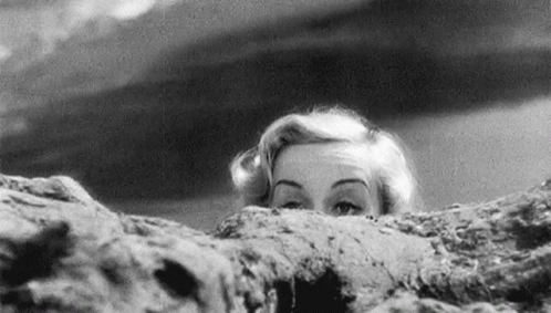 A Black And White Photo Of A Woman Peeking Over A Rock . GIF