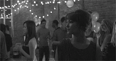 A Black And White Photo Of A Woman Standing In A Crowded Room At A Party . GIF