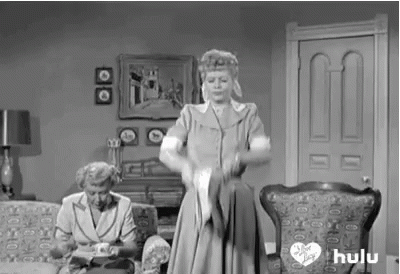 A Black And White Photo Of A Woman Standing In A Living Room Holding A Cat . GIF