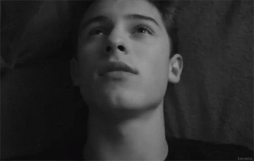 A Black And White Photo Of A Young Man Laying On A Bed Looking Up At The Sky . GIF