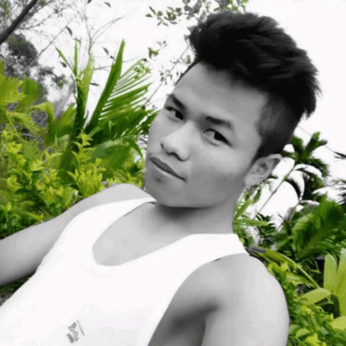 A Black And White Photo Of A Young Man Wearing A White Tank Top With A Few Plants Behind Him GIF
