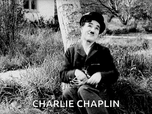 A Black And White Photo Of Charlie Chaplin Sitting Next To A Tree GIF