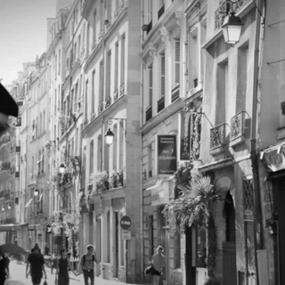 A Black And White Photo Of People Walking Down A Narrow Street With A Sign That Says Hotel Voyageur On It GIF