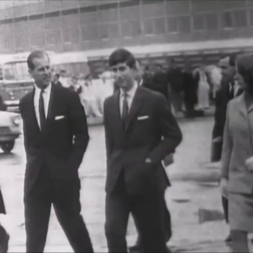 A Black And White Photo Of Two Men In Suits Standing Next To Each Other On A Street . GIF
