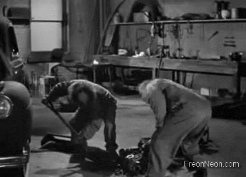 A Black And White Photo Of Two Men Working On A Car Engine In A Garage . GIF
