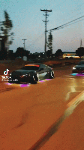 A Black Car Is Driving Down A Dirt Road With A School Bus In The Background .. GIF