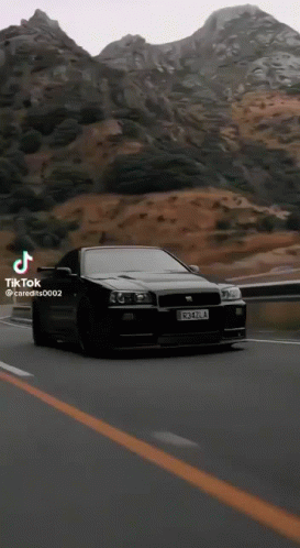 A Black Car Is Driving Down A Road In Front Of Mountains . GIF