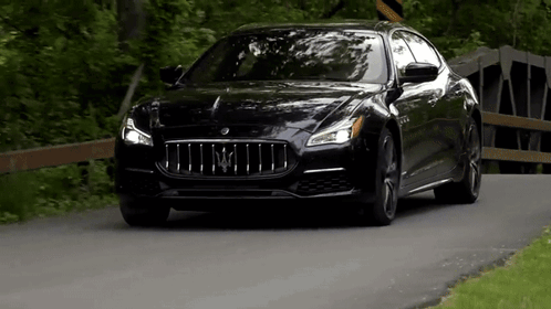 A Black Maserati Is Driving Down A Road Next To Trees . GIF